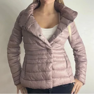 Rosa dunjacka - Snygg rosa dunjacka med hög krage och knappar. Jackan har en figurnära passform och långa ärmar. Perfekt för kyligare dagar.