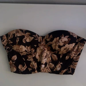 Blommig axelbandslös topp - Snygg axelbandslös topp med blommigt mönster i beige och svart. Perfekt för en somrig look. Toppen har en korsettliknande design som ger en fin passform.