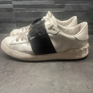 Valentino sneakers i vitt och svart - Snygga Valentino sneakers i vitt med svart band över snörningen. Skorna har en unik design med nitar runt hälen och en robust sula. Perfekta för en stilren look. Dom är använda så Finns slitage. Men själva skon är fortfarande fin. Storlek 44! Box ingår ej.