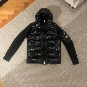 Svart pufferjacka från Moncler - Snygg svart pufferjacka från Moncler med glansigt material och huva. Jackan har långa ärmar och dragkedja framtill. Det finns en ficka med knapp på ärmen och Moncler-loggan syns tydligt. Perfekt för kyligare dagar.