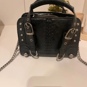 Svart handväska med detaljer - Svart handväska med coola detaljer. Har använt många gånger och tecken på användning syns, metallen har blivit lite rustigt men annars har väskan tagits bra hand om.  Kedjan har knyts ihop (bild 3)och då kan den se kortare ut om man ska ha crossbody. Skriv om du har funderingar💕💕