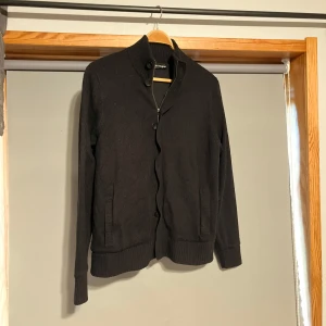 Cardigan jacka - Snygg svart stickad jacka med dragkedja och knappar. Den har en hög krage och ribbade muddar vid ärmslut och nederkant. Perfekt för kyligare dagar.