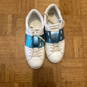 Valentino open - Snygga Valentino sneakers i vitt med en iögonfallande blå detalj över snörningen. Skorna har en klassisk design med rund tå och är tillverkade i skinn. Perfekta för en stilren look.