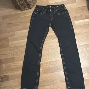 Svarta jeansbyxor från True religion med guld stitching - Snygga svarta jeansbyxor från True Religion med kontrasterande sömmar. Byxorna har en straight fit och klassisk femficksdesign. Perfekta för en stilren look.