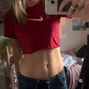 Röd croppad t-shirt från Nike - Säljer en röd croppad t-shirt från Nike med korta ärmar och det klassiska swoosh-loggan i vitt på bröstet. Perfekt för en sportig och avslappnad stil. Passar bra till jeans eller träningskläder.