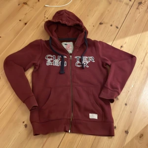 Vinröd hoodie från Cutter & Buck - Säljer en snygg vinröd hoodie från Cutter & Buck med dragkedja och tryck framtill. Bra skick på tröjan men är det är ett helt annat märke på luvan man märker inte det på.annars väldigt fint sick.Märket är känt för sin kvalitet och stil.