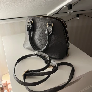 Mörk grå handväska med dragkedja - Snygg svart handväska i skinn med guldfärgade detaljer och dragkedja. Väskan har både handtag och en avtagbar axelrem för flexibilitet. Insidan är rymlig med ett mönstrat foder. 