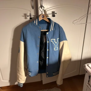 Blå och vit bomberjacka av äkta leder med bokstavsdetalj - Snygg bomberjacka i blått och vitt med en stor bokstav 'Y' på bröstet och siffran '59' på ärmen. Jackan har vita ärmar och blå kropp, med ribbade muddar och krage i randigt mönster. Perfekt för en cool och avslappnad stil. Jackan är äkta leder och i bra skick!