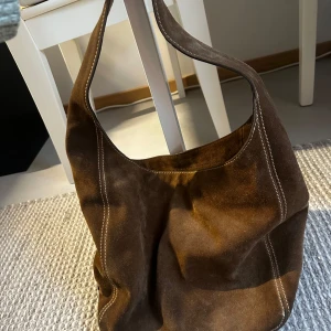Brun axelväska från Michael Kors - Snygg brun axelväska i mocka från Michael Kors. Väskan har en praktisk ficka på utsidan i skinn och en dragkedja på insidan för säker förvaring. Perfekt för att bära med sig det nödvändigaste med stil.