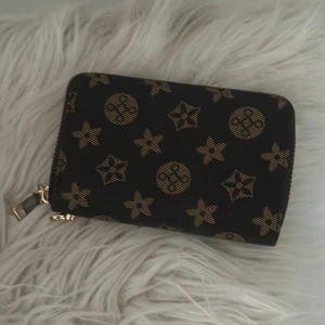 Svart och guld plånbok med dragkedja - Snygg svart plånbok med guldmönster och dragkedja. Perfekt för att hålla ordning på kort och kontanter. Den har flera fack inuti för enkel organisering.