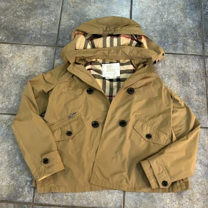 Beige Burberry jacka dam  - Säljer en snygg beige jacka från Burberry med klassiskt rutigt foder. Jackan har en avtagbar huva och knappar framtill. Perfekt för höst och vår! Köp från johnells äkta