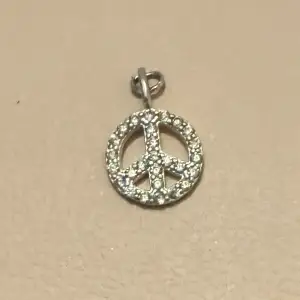 Snyggt och stilrent hänge i form av en peace-symbol. Det är silverfärgat och dekorerat med små glittrande stenar som ger en extra touch av elegans. Perfekt för att sätta på ett halsband och bära till både vardags och fest.