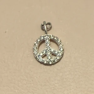 Silverfärgat peace-hänge - Snyggt och stilrent hänge i form av en peace-symbol. Det är silverfärgat och dekorerat med små glittrande stenar som ger en extra touch av elegans. Perfekt för att sätta på ett halsband och bära till både vardags och fest.