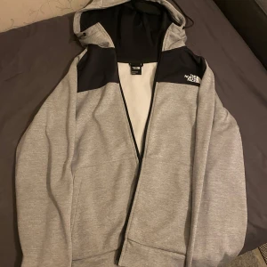 Grå hoodie från The North Face - Säljer en grå hoodie från The North Face i mycket bra skick. Den har en dragkedja framtill och en justerbar huva. Perfekt för höst och vår, med en bekväm passform. Svarta detaljer på axlarna ger en snygg kontrast. Perfekt för både vardag och träning!