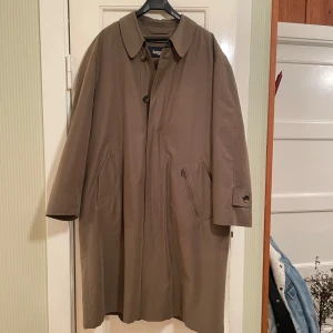 Mullvadsfärgad varmfodrad herrock från Bugatti - Säljer en 80-tals mörkbeige herrock från Bugatti i storlek 46. Den har en stilren design med knappar och en rutig/svart varmfodrad insida. Fodret löstagbart. Perfekt över kostymen höst och vinter. Utsida borstad poplin. Två öppna framfickor och en med dragkedja, innerficka med dragkedja. Ett måste för den stilmedvetnes garderob!