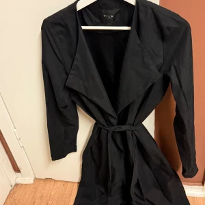 Svart trenchcoat från VILA - Snygg svart trenchcoat från VILA i storlek S. Jackan har en klassisk design med knytband i midjan och långa ärmar. Perfekt för höst och vår, och funkar både till vardags och fest. 🖤