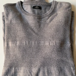 Grå tröja från Marks & Spencer - Säljer en stilren grå tröja från Mats Norgaard Danmark.  Den har ett diskret mönster över bröstet och är tillverkad i ett mjukt material. Perfekt för både vardag och lite finare tillfällen. Tröjan har långa ärmar och en rund halsringning.