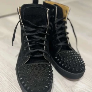 Svarta sneakers med nitar - Säljer ett par svarta sneakers med coola nitar på tån och sidorna. De har en röd sula som ger en snygg kontrast. Skorna är i mycket bra skick och har snörning för perfekt passform. Perfekta för att ge din outfit en edgy touch! En gång använda pris kan diskuteras.