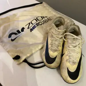 Säljer ett par Nike Zoom Mercurial fotbollsskor i beige och svart. Skorna har snörning och är i bra skick, perfekta för fotbollsträning. De kommer med en praktisk förvaringspåse. Skorna har en snygg design med svarta detaljer och är gjorda för att ge bra grepp på planen. Pris kan diskuteras vid snabb affär