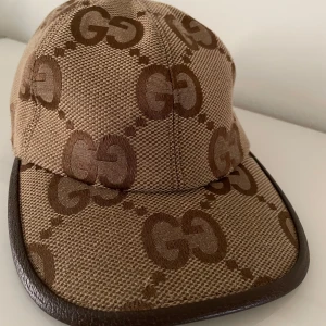 Beige keps från Gucci - Säljer en snygg beige keps från Gucci med det klassiska GG-mönstret i brunt. Kepsen har en stilren design med en böjd skärm och är perfekt för både vardag och fest. Den är i mycket bra skick och har en bekväm passform. Perfekt accessoar för att lyfta din outfit!