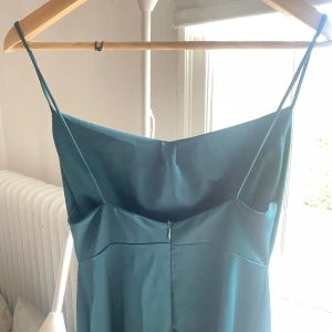 Turkos långklänning med smala axelband och öppen rygg! - Säljer en elegant turkos långklänning med smala axelband. Perfekt för sommarens fester eller en fin middag. Klänningen har en enkel och stilren design med en lätt glansig yta.