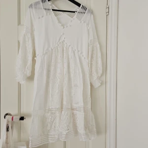 Vit spetsklänning från JardhVue - Säljer en superfin vit klänning från JardhVue. Den har ett vackert spetsmönster och volanger som ger en romantisk känsla. Klänningen är långärmad och har en v-ringad design med små söta bollar längs kanten. Perfekt för sommarens fester eller semester