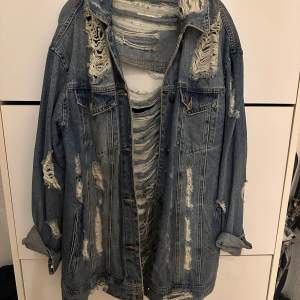 Säljer en riktigt cool och sliten jeansjacka i blått. Den har en oversized passform och är full av slitningar både fram och bak, vilket ger den en grunge-stil. Perfekt för höst och vår när du vill ha en avslappnad look. Jackan har långa ärmar och klassisk krage.