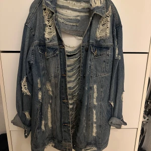 Sliten jeansjacka i blått - Säljer en riktigt cool och sliten jeansjacka i blått. Den har en oversized passform och är full av slitningar både fram och bak, vilket ger den en grunge-stil. Perfekt för höst och vår när du vill ha en avslappnad look. Jackan har långa ärmar och klassisk krage.