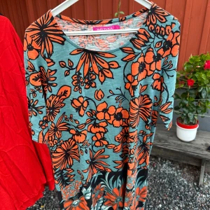 Blommig klänning från Indiska - Säljer en superfin klänning/tunika från Indiska i storlek M. Den har ett färgglatt blommönster i orange och svart på en ljusblå bakgrund. Klänningen är kortärmad och perfekt för sommarens alla fester. 