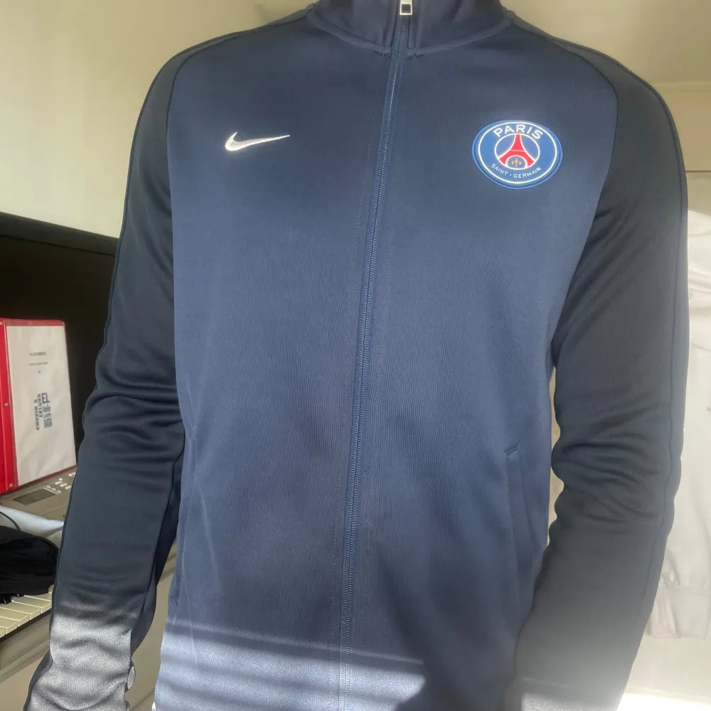 Säljer en snygg mörkblå träningsjacka från Nike med PSG-logga på bröstet. Jackan har en dragkedja framtill och långa ärmar. Assnygg och i perfekt skick, inga defekter. Ganska svår att få tag på idag. . Jackor.