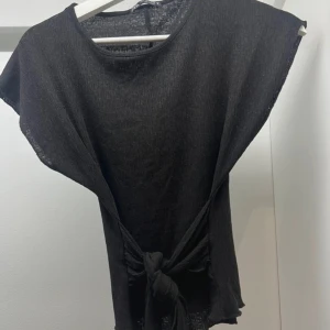 Svart topp med knyt - Säljer en svart topp med korta ärmar och knytning i midjan. Toppen har en enkel och stilren design, perfekt för både vardag och fest. Materialet ser ut att vara lätt och luftigt.