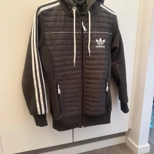 Svart quiltad jacka från Adidas - Säljer en snygg svart jacka från Adidas. Den har en dragkedja framtill och vita detaljer som de klassiska Adidas-ränderna på ärmarna. Jackan har en huva med vita snören och två fickor med dragkedja. Perfekt för höst och vår! XS