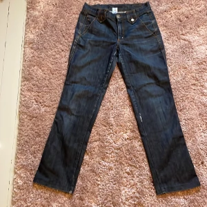 Mörkblå jeans med detaljer - Säljer ett par snygga low/midwaist Marc Jacobs jeans med detaljer som knappar och en liten bältesdetalj bak. Älskar de men passar inte riktigt mig. Den högra knapp-detaljen där bak är lite trasig men löses lätt med en ny knapp eller säkerhetsnål.