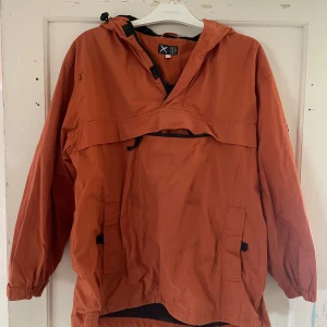 Orange vindjacka från Cross - Säljer en cool orange vindjacka från Cross. Den har en luva och praktiska fickor framtill. Perfekt för utomhusaktiviteter med ett tryck av en cyklist på baksidan. Jackan är i bra skick och redo för nya äventyr!