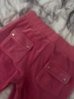 Rosa velourbyxor Juicy Couture - Säljer ett par klassiska rosa mjukisbyxor från Juicy Couture i mjuk velour. Byxorna har bred resår i midjan med dragsko, stora fickor med lock och silverfärgade knappar bak samt broderad logga framtill. Perfekta för en chill och trendig look. De har en liten defekt på byxan som är franslim men går säkert att få bort lätt, har bara inte försökt. Inget som man märker väl heller. 