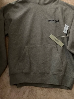Grå Essentials Fear of God hoodie - Priset är inte sats det kan förhandlas. Snygg grå hoodie från Essentials Fear of God med svart logga på bröstet. Klassisk känguruficka framtill och ribbade muddar. Hoodien har huva och är tillverkad i mjukt bomullsmaterial. Perfekt för en avslappnad streetwear-look.