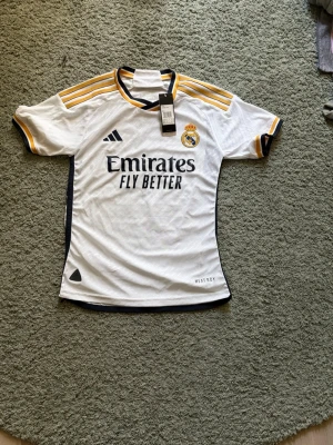 Real Madrid matchtröja Bellingham - Vit Real Madrid matchtröja från Adidas med Bellingham och nummer 5 på ryggen. Tröjan har gula och svarta detaljer, klubbmärke på bröstet och 'Emirates Fly Better' tryckt framtill. Korta ärmar och sportig passform, perfekt för fotbollsträning eller match.