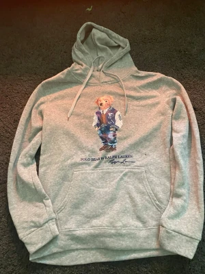 Grå hoodie Polo Bear Ralph Lauren - Snygg grå hoodie från Ralph Lauren med den ikoniska Polo Bear-trycket framtill. Hoodien har huva med dragsko och en stor magficka. Tillverkad i mjukt bomullsmaterial, perfekt för en avslappnad och trendig stil.