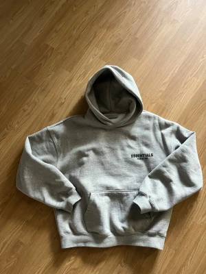 Grå Essentials Fear of God hoodie - Säljer en grå hoodie från Essentials Fear of God med broderad logga på bröstet. Hoodien har en stor huva, magficka och ribbade muddar vid ärmar och nederkant. Tillverkad i mjukt bomullsmaterial som är riktigt skönt att ha på sig.