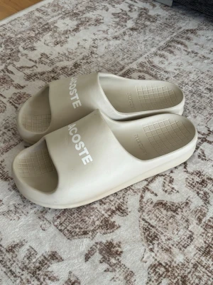 Beige tofflor från Lacoste - Snygga beige tofflor från Lacoste med vit logga på ovansidan. Tillverkade i ett mjukt och bekvämt gummimaterial med rutmönstrad sula för extra grepp. Perfekta att glida i hemma eller på stranden.
