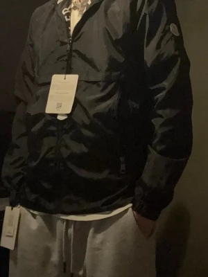 Svart vindjacka från Moncler - Snygg svart vindjacka från Moncler med dragkedja framtill och elastiska muddar vid ärmsluten. Jackan har Moncler-logga på ärmen och praktiska sidofickor med dragkedja. Tillverkad i lätt och slitstarkt material, perfekt för blåsiga dagar.