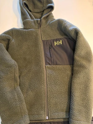 Olivgrön teddyjacka Helly Hansen - Mysig olivgrön teddyjacka från Helly Hansen med huva och hel dragkedja. Jackan har en bröstficka i kontrasterande tyg med HH-logga och dragkedja. Tillverkad i fluffigt teddy-material som håller dig varm och bekväm. Perfekt för kyliga dagar.
