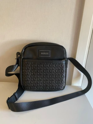 Svart axelväska från Guess - Snygg svart axelväska från Guess med justerbar axelrem. Väskan har ett präglat mönster med Guess-loggor på framsidan och en stilren logopatch upptill. Tillverkad i konstläder med en fyrkantig form och praktiskt ytterfack.