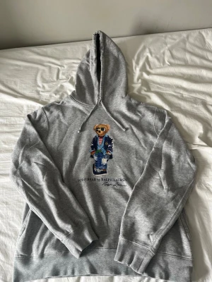 Grå hoodie Polo Bear Ralph Lauren - Säljer en grå hoodie från Ralph Lauren med den ikoniska Polo Bear-trycket på bröstet. Hoodien har huva och en klassisk passform. Materialet är mjukt och skönt, perfekt för chill dagar. Snygg detalj med björnen i blå outfit och randig tröja.