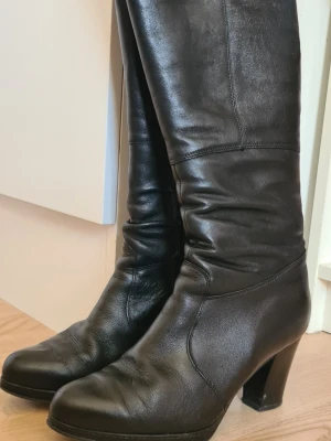 Svarta höga boots med klack - Snygga svarta boots i skinnimitation med hög klack och dragkedja på insidan. Stövlarna har en stilren design med lätt rundad tå och slät yta. Perfekta för dig som vill ha ett par klassiska boots med lite edge.