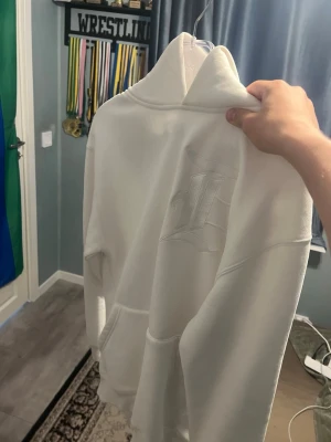 Vit hoodie med broderad logga - Säljer en stilren vit hoodie med stor broderad logga på bröstet. Hoodien har en klassisk huva och känguruficka framtill. Perfekt för dig som gillar enkel men snygg streetwear. Materialet känns mjukt och bekvämt.