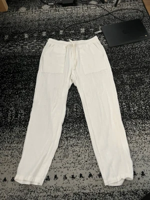 Vita linnebyxor med dragsko - Snygga vita linnebyxor med raka ben och dragsko i midjan. Byxorna har två stora fickor framtill och är tillverkade i ett lätt och luftigt linnematerial, perfekta för en avslappnad stil. Passar perfekt till sommaren och ger en fräsch look.