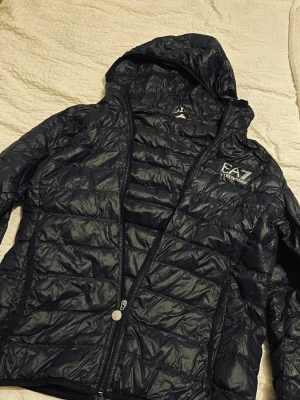Mörkblå dunjacka från Emporio Armani - Snygg mörkblå dunjacka från Emporio Armani EA7 med huva och dragkedja. Jackan har quiltad design, två sidofickor och logga på bröstet. Perfekt för kalla dagar och riktigt lätt att bära. Materialet är glansigt och ger en modern look.