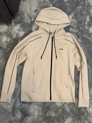 Beige hoodie från BOSS Storlek S - Snygg beige hoodie från BOSS med svarta detaljer och dragkedja framtill. Hoodien har huva med dragsko, svarta linjer längs ärmarna och diskret BOSS-logga på bröstet. Storlek S