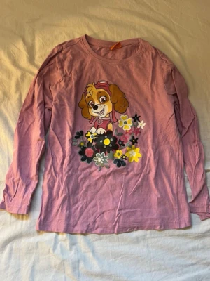 Lila långärmad topp Nickelodeon för barn  - Söt lila långärmad topp från Nickelodeon med tryck av Skye från Paw Patrol och färgglada blommor på framsidan. Perfekt för dig som gillar gulliga motiv och vill ha en bekväm och mjuk tröja i bomull.
